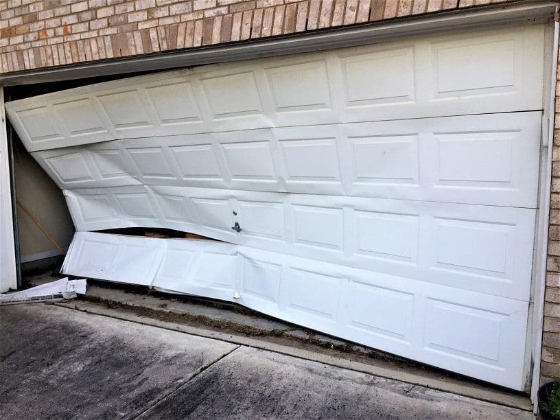 Damaged white garage door partially open, displaying bent panel and structural issues.