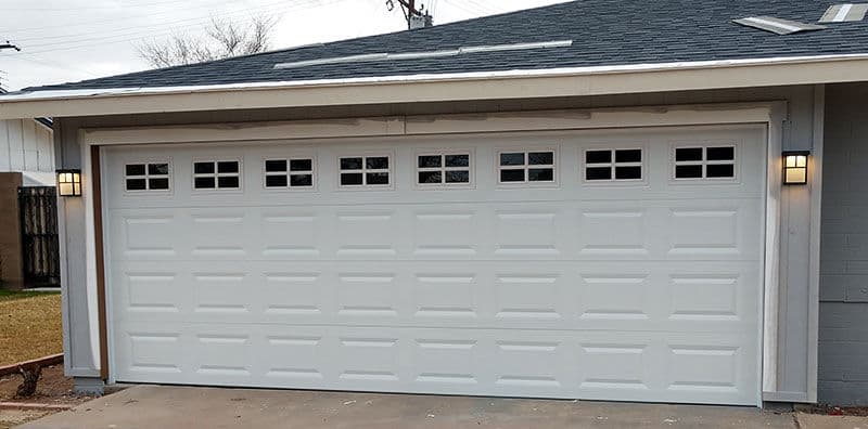 White garage door with windows, exterior lighting, and a residential home backdrop.